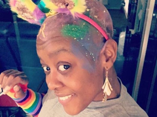 Colourful photograph of Simone with paint and glitter on her bald head, along with a headband with two rainbow coloured ears, and a unicorn's horn. Simone is smiling.
