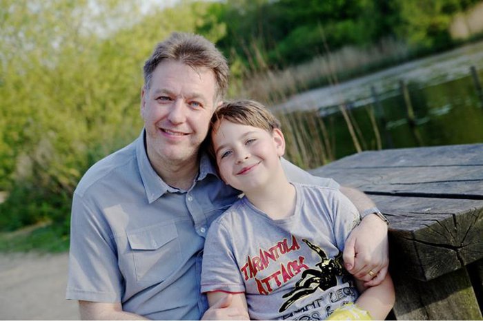 Adrian, who lives with chronic lymphocytic leukaemia (CLL), sits on a picnic table next to a lake, smiling with his child.