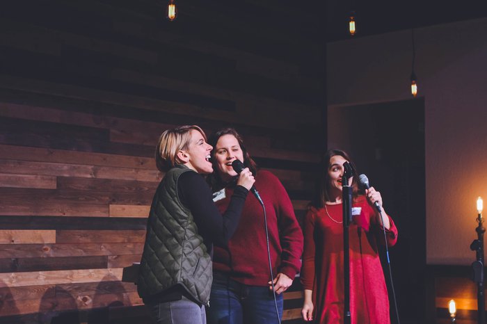 Three women laugh whilst singing karaoke on stage.