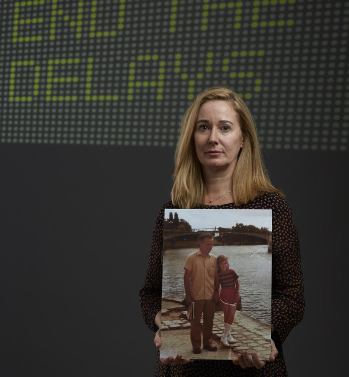 Diana Elkins stands holding a picture of her with her dad, who died of blood cancer. Behind her on the wall it says end the delays.