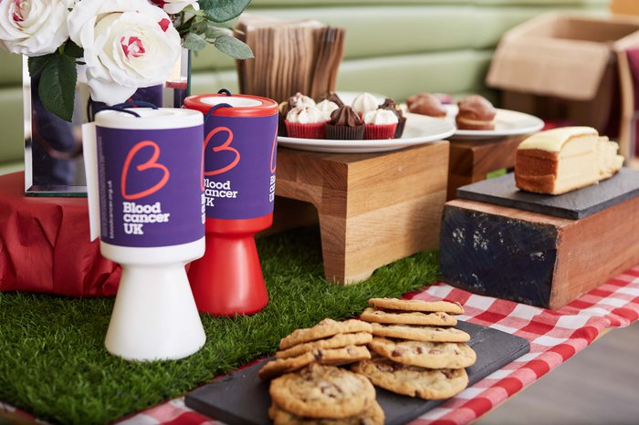 A table filled with delicious cakes and cookies, along with fundraising tins for a bake sale to raise money for Blood Cancer UK.