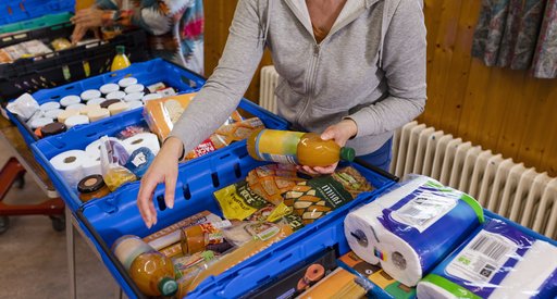 Two people are sorting through crates of food and toiletries at a food bank. There are several blue crates full of things like packets of pasta and bottles of squash.