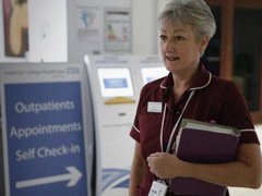 A nurse stands at an out patient clinic. She has short hair and is holding a clipboard.