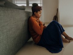 A stressed woman sits on the floor, holding her hand on her chest.