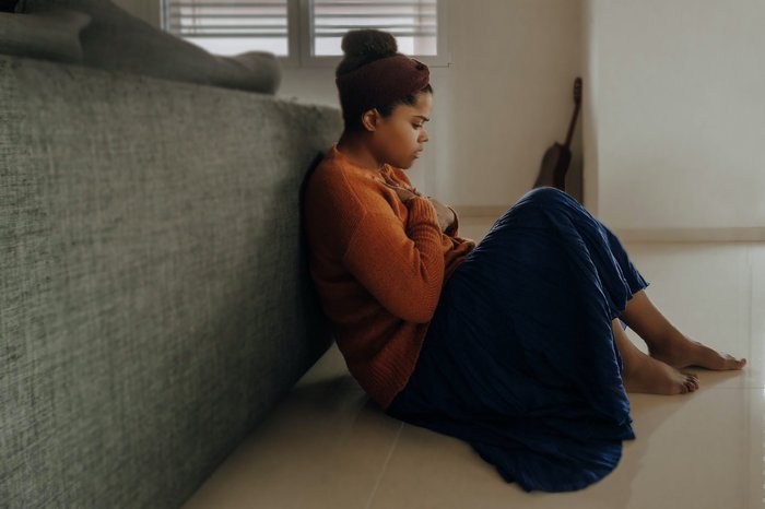 A stressed woman sits on the floor, holding her hand on her chest.