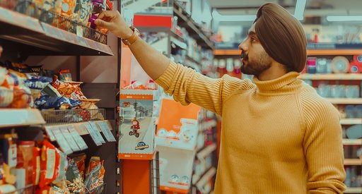 A stock image of a man wearing a turban and a yellow jumper, reaching up to grab a packaged item off a supermarket aisle.