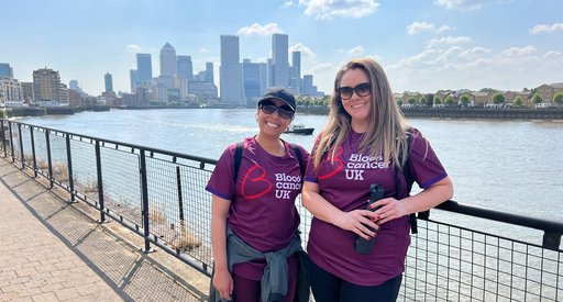 Two smiling women in Blood Cancer UK t-shirts stand next to the river Thames. The City of London is visible behind them. They are wearing sunglasses and it's a warm summer's day.