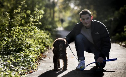 Teenage Blood Cancer UK ambassador kneeling down next to small dog on a concrete path next to bushes.