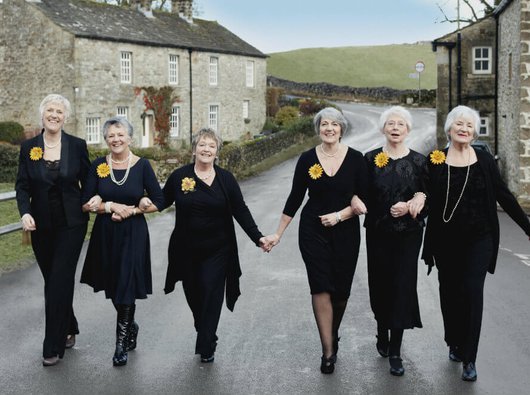 The Calendar Girls smiling and linking arms walking down a road in Yorkshire between two rows of houses.