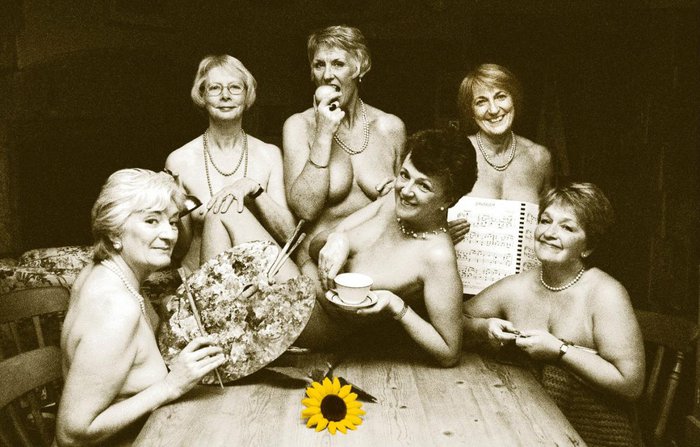 The Calendar Girls in black and white posing holding props around a wooden table and with a sunflower in colour lying on the table.