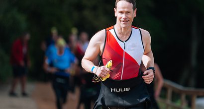 Triathlon participant smiling with wetsuit round waist and Blood Cancer UK top on whilst running.