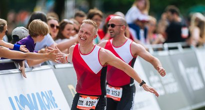 Two triathlon participants wearing Blood Cancer UK tops running and smiling whilst interacting with the crowd cheering.
