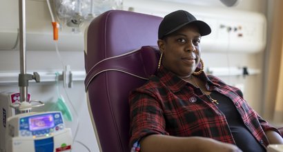 A woman sitting in a hospital receiving intravenous treatment. She's sitting in a chair, with a thin plastic tube coming out from under her rolled up shirt sleeve. There's an IV stand next to her.