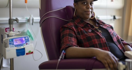 A woman sitting in a hospital receiving intravenous treatment. She's sitting in a chair, with a thin plastic tube coming out from under her rolled up shirt sleeve. There's an IV stand next to her.