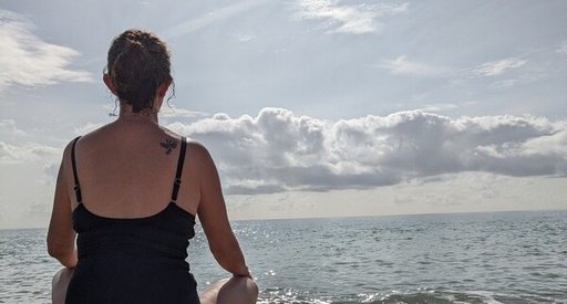 A woman in a swimming costume sitting on a pebble beach, looking out to sea.